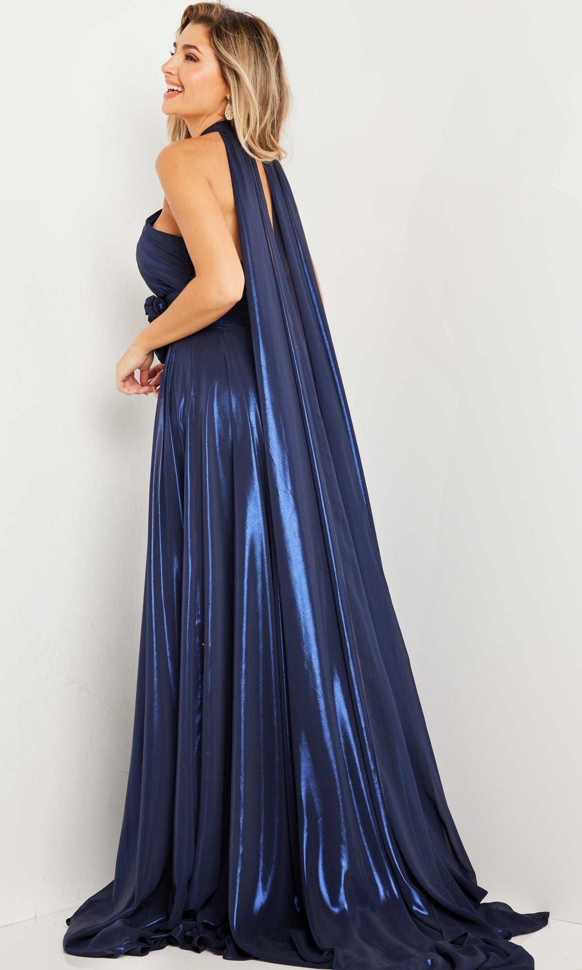A woman wearing the Long Prom Dress 37163 by Jovani, a shiny navy blue gown with a draped back, stands smiling and looking over her shoulder.